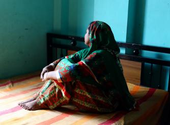 A woman sitting on a bed faces away from the camera, with her hands around her knees. 